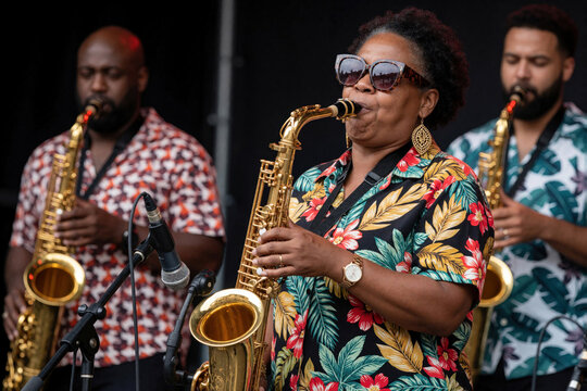 Jazz band plays saxophone music on outdoor stage. Musicians perform live concert wearing colorful shirts. People enjoy melody, rhythm, improvisation at event.