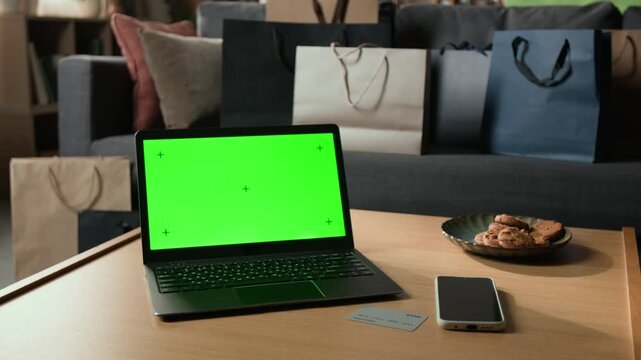 Laptop displaying green screen chroma key mockup standing on table near credit card and smartphone with shopping bags. Footage suitable for e-commerce retail ads and digital payment promotions
