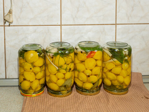 Four Jars of Preserved Yellow Pattypan Squash. Four glass jars filled with small yellow pattypan squash are displayed side by side against a tiled kitchen background. The jars contain preserved vegeta