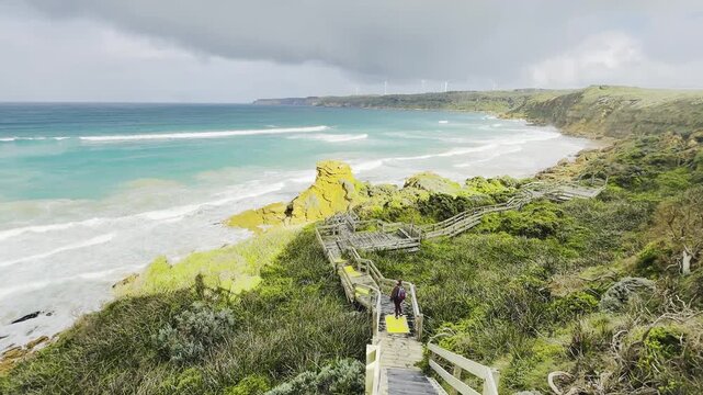A girl traveler stands on a wooden boardwalk, admiring the stunning rock formations at the beach, Yellow Rock, Portland, Victoria, Australia.