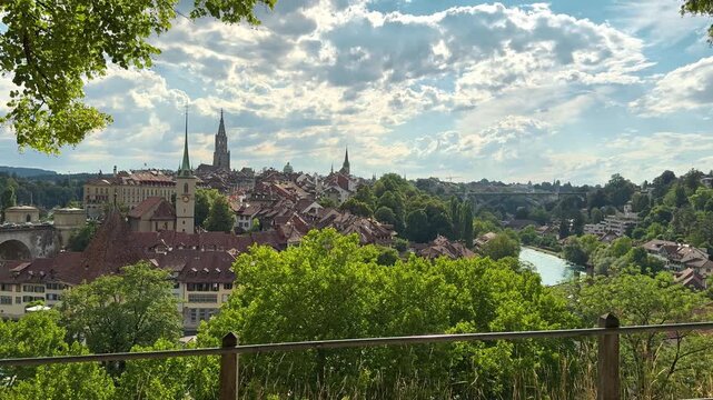 Bern old town rises above trees beside the winding Aare River in clear daylight, Historic towers rooftops and bridges create a scenic Swiss capital panorama