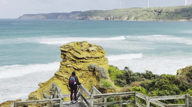 A girl traveler stands on a wooden boardwalk, admiring the stunning rock formations at the beach, Yellow Rock, Portland, Victoria, Australia.