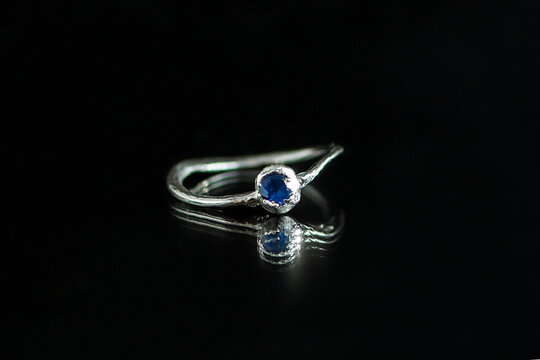 A silver ring with a blue gemstone sits on a reflective black surface. The ring has a simple design, showcasing the vibrant color of the stone.