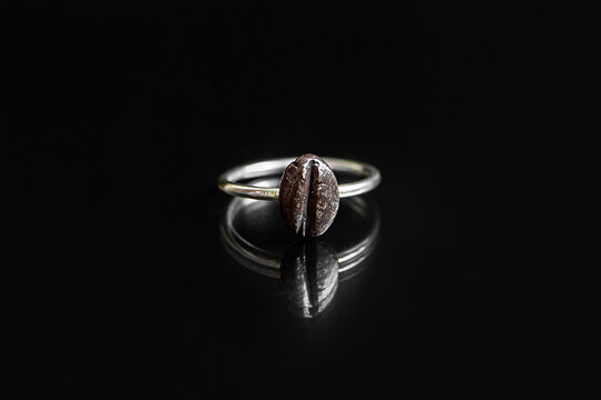 A silver ring featuring a coffee bean design, placed on a reflective black surface. The ring showcases intricate details of the coffee bean.
