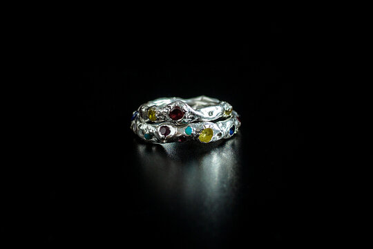 A close-up of a silver ring adorned with colorful gemstones on a dark background. The ring features intricate designs and a polished finish.