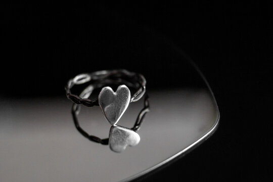 A silver ring with two heart shapes intertwined, resting on a reflective surface. The background is dark, emphasizing the ring's design and shine.