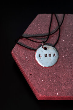 A silver pendant with the word LUNA engraved on it, hanging from a black cord. The pendant is placed on a textured red hexagonal surface.