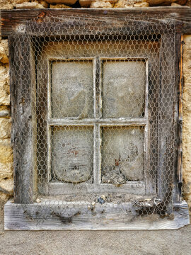 Dilapidated wooden window frame with chicken wire mesh and torn fabric panes in an old stone building wall during daylight