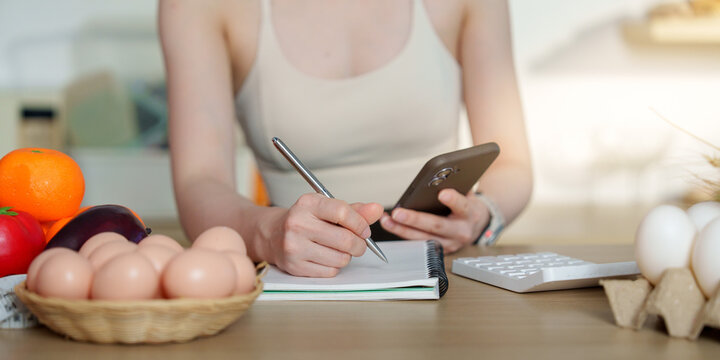 Cropped hands writing meal plan in notebook near fresh eggs and fruits for nutrition tracking