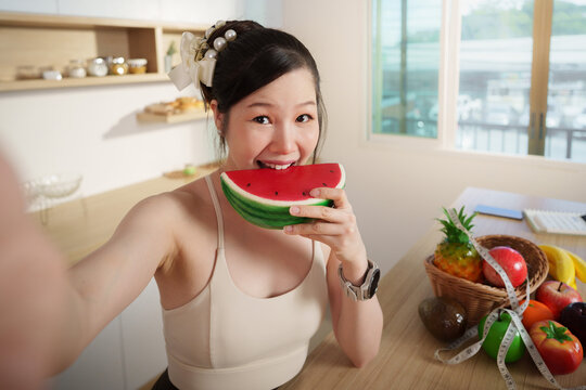 Smiling Asian woman taking selfie while eating fresh watermelon slice in kitchen for healthy lifestyle