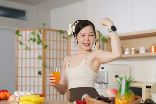 Fit Asian woman flexing bicep muscle and holding fresh orange juice in kitchen for healthy lifestyle
