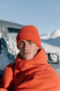 Outdoor winter portrait of a young man in an orange puffer jacket and beanie leaning by a snowy car for seasonal lifestyle background and banner design