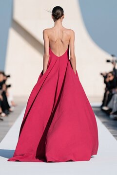 Slim model in backless fuchsia gown walking on runway with architectural backdrop for fashion show poster and editorial layout