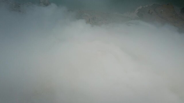 Dense volcanic gas drifts over yellow sulfur vents beside the turquoise crater lake as the drone flies above the Ijen volcano site.