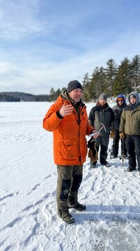 An experienced outdoor guide passionately instructs a group of enthusiastic anglers on a vast frozen lake surrounded by a pristine winter snowscape
