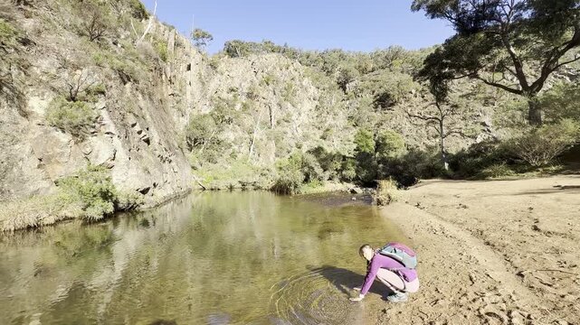 A girl hiker rests by the Werribee River, touching the water, during the challenging Werribee Gorge Circuit Walk - one of the best trails near Melbourne, Victoria, Australia.