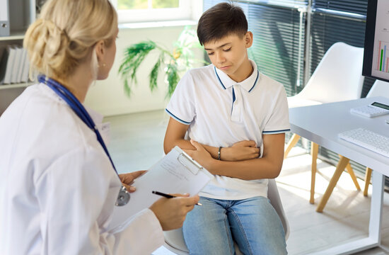 Doctor examining boy patient with abdominal pain. Pediatrician notes symptoms in modern clinic office. Teen seeks diagnosis and care during consultation. Clear concept of child healthcare.
