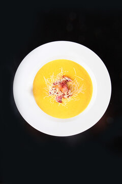 Creamy yellow soup in white bowl. Crispy bacon and grated cheese garnish. Black background highlights dish. Gourmet appetizer. Top view