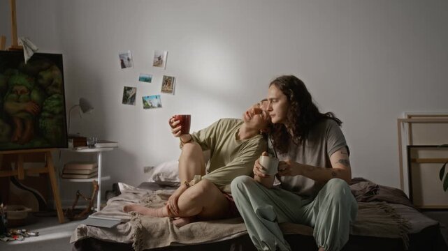 Caucasian young couple drinking coffee together on bed and cuddling affectionately in wide shot. Footage suits lifestyle advertising, relationship storytelling, and domestic wellness campaigns