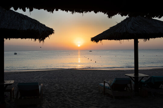 Tropical beach sunset. Thatched umbrellas over lounge chairs on sand. Calm sea, sun over horizon, boats, birds. Peaceful vacation
