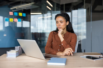 Young businesswoman with dark hair and brown shirt sitting at a desk, concentrating on a laptop...