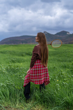 Young woman holding vintage badminton racket stands in green field under dramatic cloudy sky