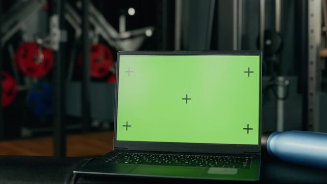 Laptop displaying chroma key green screen with tracking markers resting in gym with camera zooming in. Suitable for fitness app UI mockup and online training platform presentation