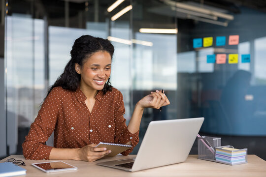 Young businesswoman smiling at her laptop while taking notes and joining a virtual meeting, planning projects and staying productive in a modern office workspace