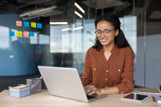 Young professional woman with glasses smiling, typing on a laptop computer at a desk in a contemporary office setting, focusing on digital work and business productivity