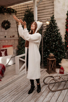 Festive mood captured as smiling young woman in white cardigan catches falling snow on decorated porch