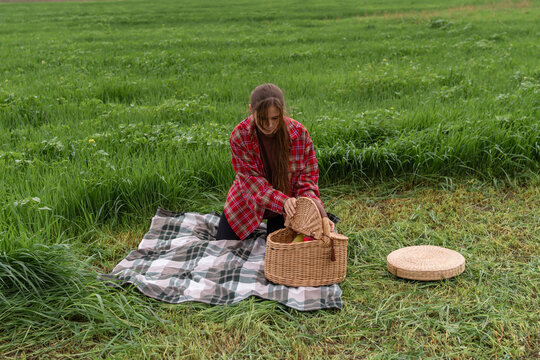 Woman opening wicker basket on plaid blanket in lush green meadow grass