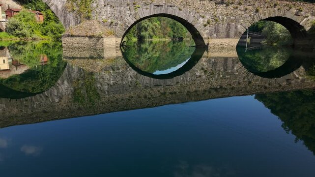 Aerial drone flying under the famous Ponte della Maddalena, also known as the Devil's Bridge, reflecting on the Serchio River in Tuscany