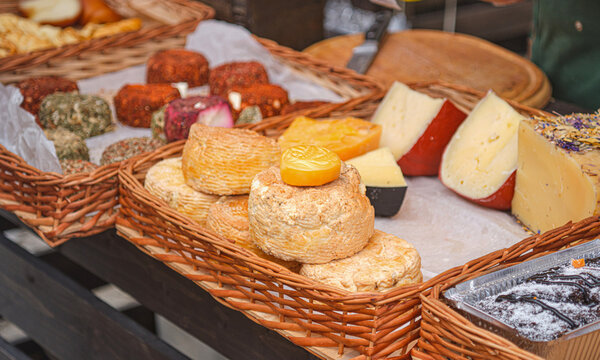 Assortment of artisan craft cheeses displayed in wicker baskets at an outdoor farmers market counter. Assorted goat and cow milk cheeses with herbs, spices, and crusts