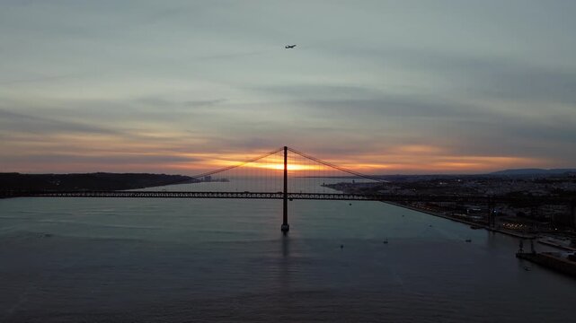 Panoramic drone shot of 25 de Abril Bridge, Lisbon at sunset. Golden hour sky, clouds. Airplane takes off in background. Portugal
