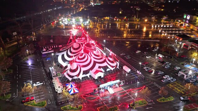 A drone pans right across a black-and-white "horror circus" tent featuring red lights and marked parking spaces for vehicles; the scene is set at a mall with some vehicles driving by, located in the S