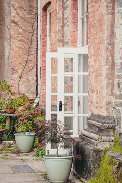Vertical image of an inviting and tranquil rustic courtyard featuring open white french doors leaning against an old brick wall, surrounded by potted plants and mossy stone elements