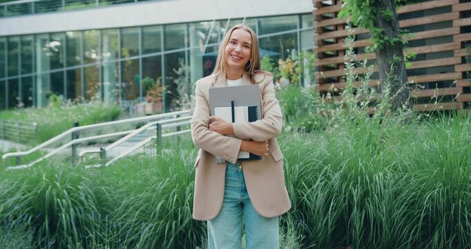 Professional young woman with long hair holding a laptop and standing in a trendy coworking space, looking at the camera with a friendly smile, outdoors