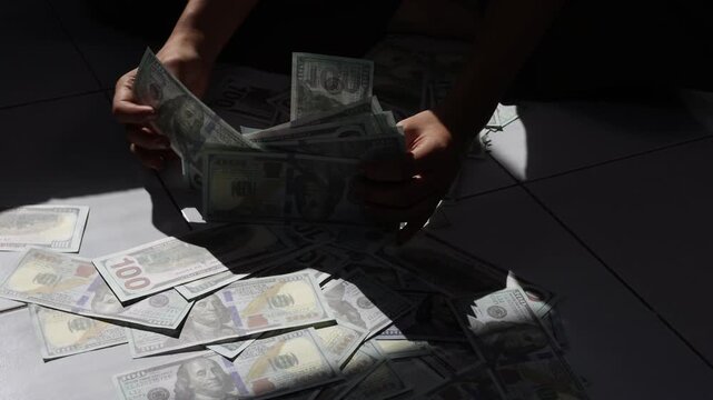 Person playing with a pile of one hundred dollar bills. Hands are grabbing and arranging a large amount of us one hundred dollar bills on a floor.