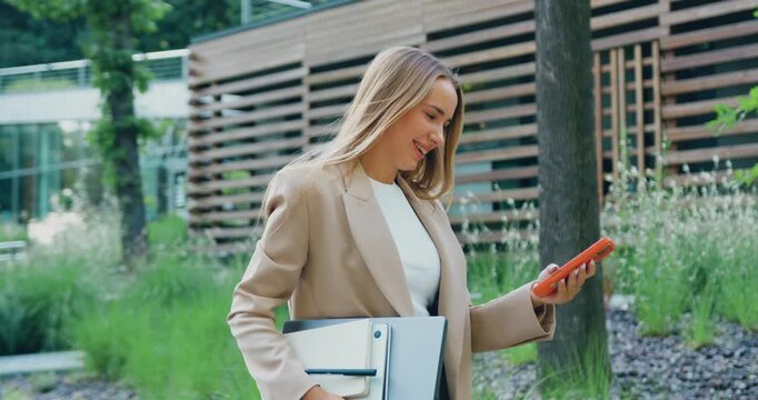 Side view of young Caucasian businesswoman in using smartphone and holding laptop while walking in front of modern building at city park. Business and people concept.
