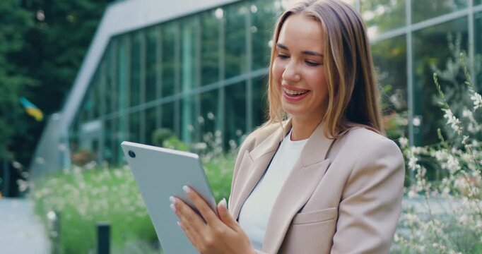 Portrait of young woman in city park in suit, working on tablet on commercial glasses building background, outdoors