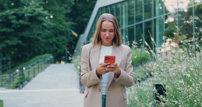 A beautiful young woman walks by a commercial building while texting on her smartphone and holding laptop yet professional appearance. Urban background. Modern lifestyle, freelance or business concept