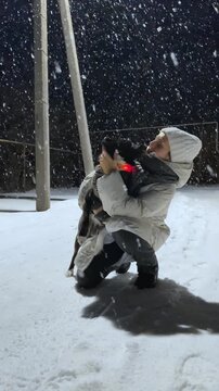 On a winter night, under a streetlamp, in a heavy snowfall, a young, happy woman is hugging and kissing her dog; the dog is wearing a glowing collar for nighttime walks.