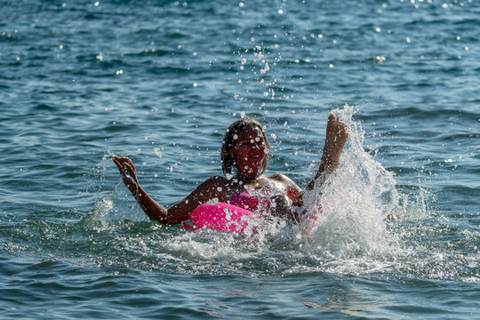 Woman splashing water joyfully while floating on pink inflatable ring in sea during summer vacation