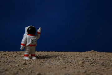 A miniature astronaut toy standing on a rocky, sandy surface, raising one hand against a deep blue background.  © Andi