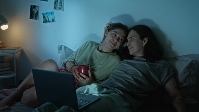 Young adult couple watching streaming video on laptop computer while resting in bed together. Perfect for online streaming platform marketing, modern lifestyle commercials and tech branding