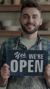 Portrait of joyful young man raising Yes we are open banner advertising pottery workshop developing small business. Traditional art studio and advertisement concept.