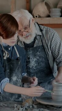 Skillful boy decorating ceramic pot under guidance of caring grandpa in arts studio. People using pottery wheel and traditional tools.