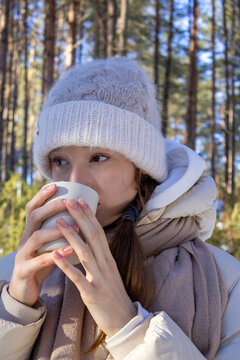 Teenage girl drinks hot beverage from cup with focused expression during winter forest outing. Concept of cozy warmth and comforting hot drink ritual during cold winter outdoor adventure.