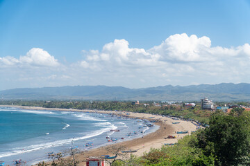 Pangandaran beach with golden sands curve along turquoise waves. White clouds drift above distant, hazy mountains. Beachgoers relax, swim, or stroll near boats. Blue sky contrasts with green foliage. © Rizky Rahmat Hidayat