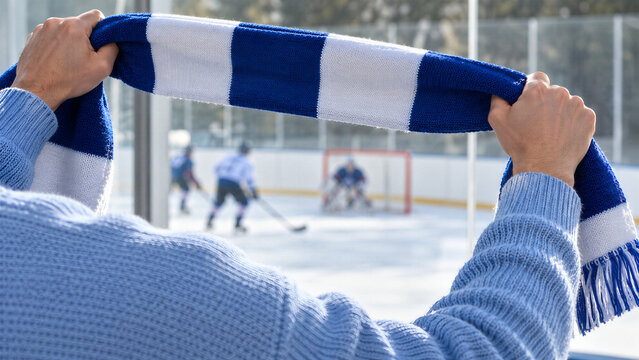 Male fan holding blue and white scarf watching ice hockey game from stands. Blurred hockey players and goalie on rink behind. Winter sport fan support and team spirit concept.